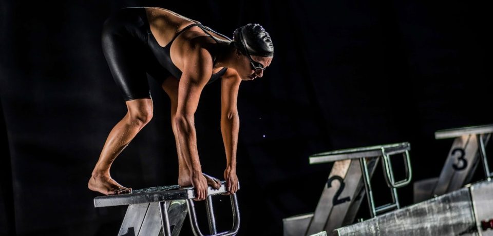 Optimised alt text: Female competitive swimmer in black swimsuit, goggles, and cap, poised on starting block ready for race in indoor pool, showcasing sports and athletic preparation.