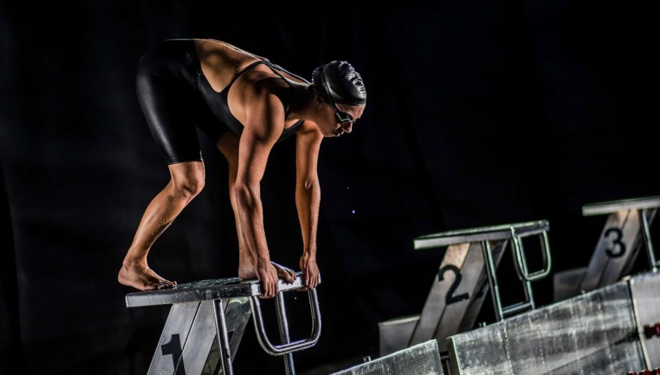 Optimised alt text: Female competitive swimmer in black swimsuit, goggles, and cap, poised on starting block ready for race in indoor pool, showcasing sports and athletic preparation.
