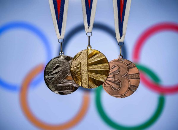 Gold, silver, and bronze medals hanging against a blurred Olympic rings backdrop, representing sporting success and victory.