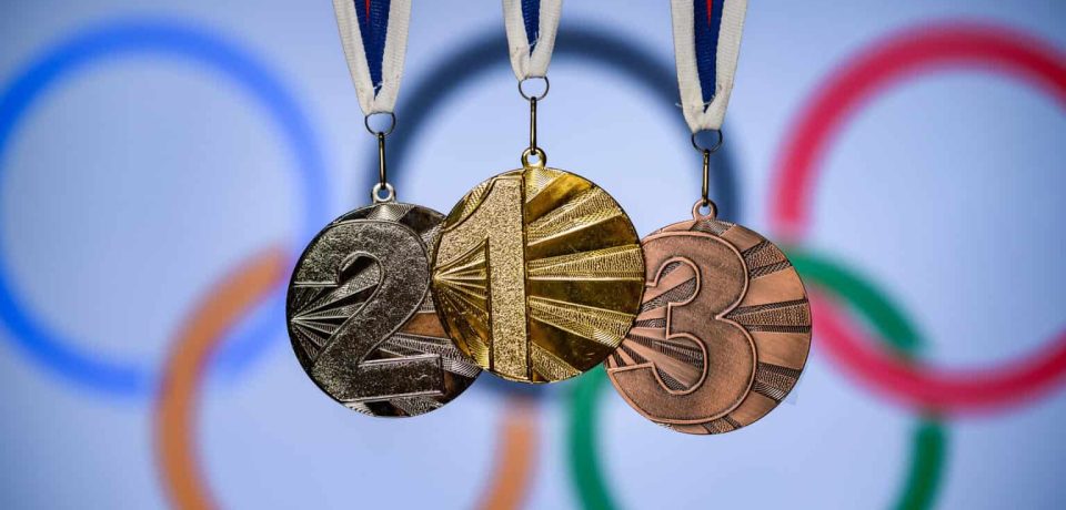 Gold, silver, and bronze medals hanging against a blurred Olympic rings backdrop, representing sporting success and victory.