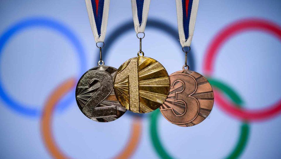 Olympic medals with the Olympic rings in the background, symbolising achievement and excellence in sports. Gold, silver, and bronze medals hanging against a blurred Olympic rings backdrop, representing sporting success and victory.